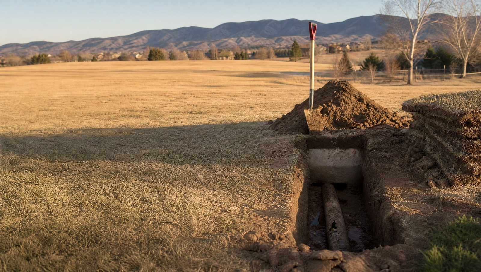 Hand dug excavation exposing a cracked septic line pipe, with a shovel standing in the spoil pile