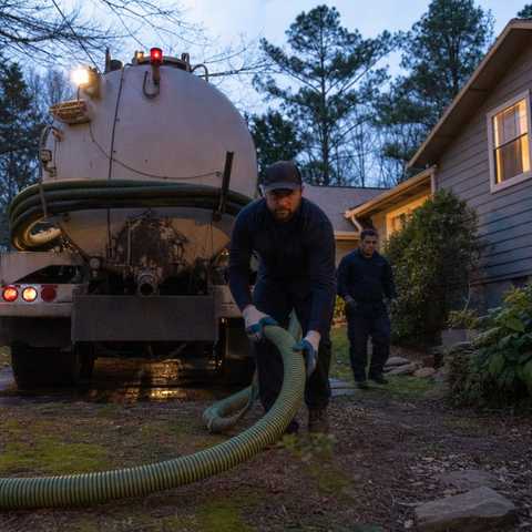 Septic pump truck at a home at dusk with a crew pulling hose for emergency service